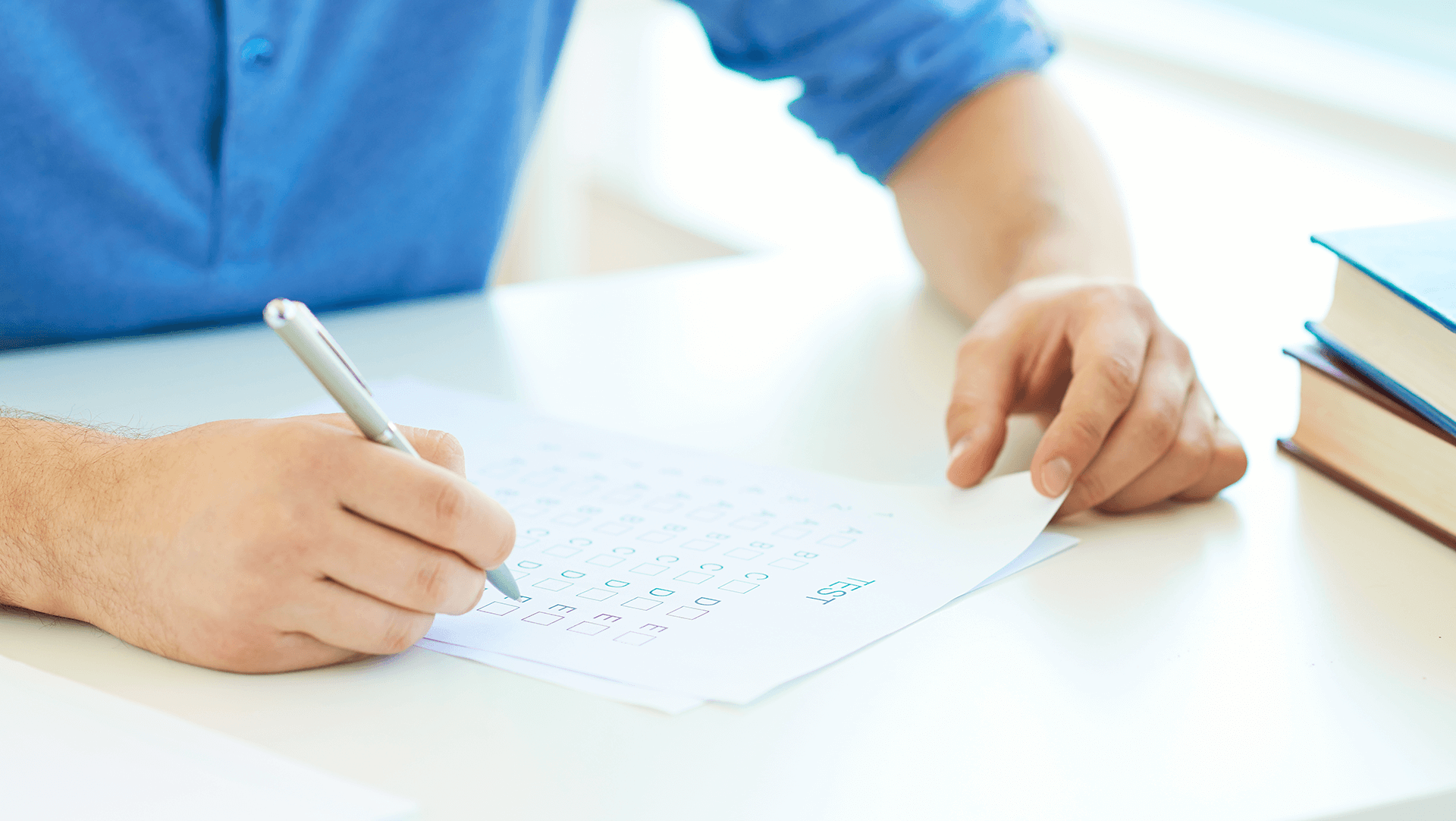 A person in a blue shirt filling out a questionnaire or survey on a paper with a pen, sitting at a white desk with books to the side.