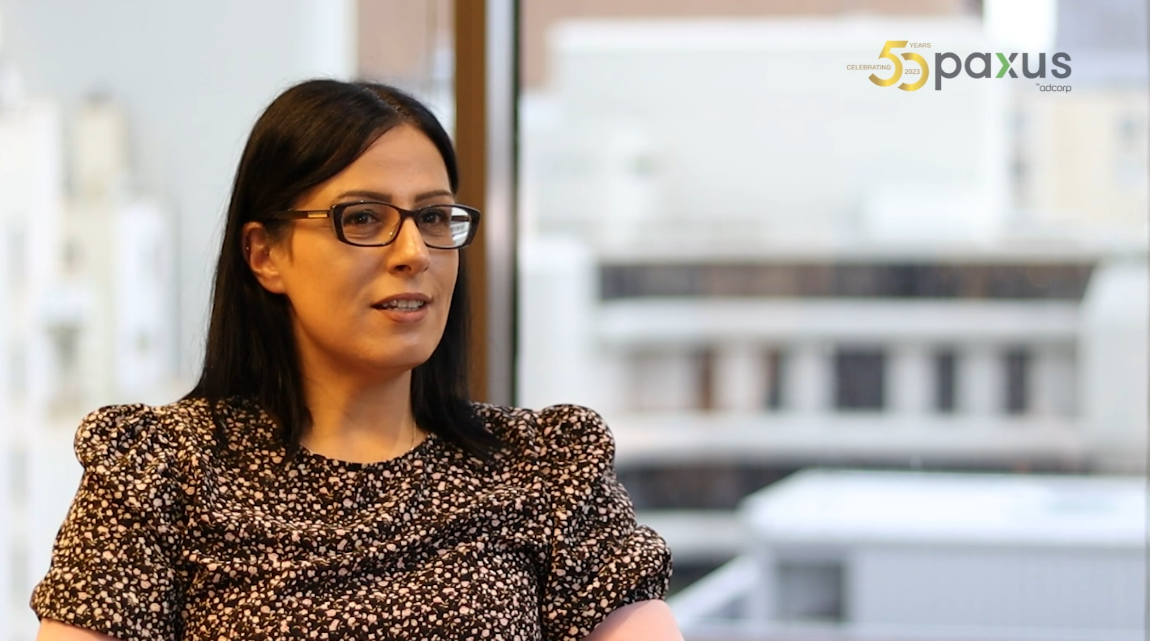 A professional woman in a patterned blouse sits engaged in an interview with a blurred office background, showcasing a workplace environment.