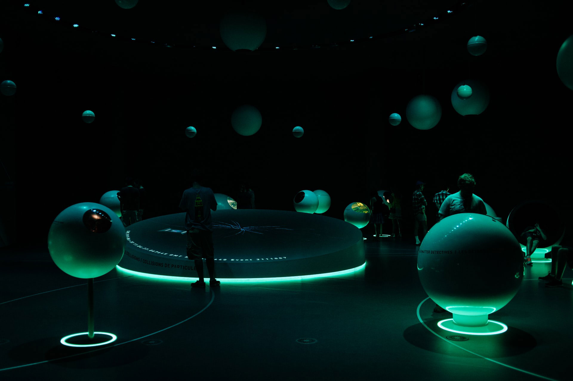 A dimly lit room with glowing green spherical displays and circular installations. People are visible interacting with the exhibits. The ambiance is futuristic and immersive.