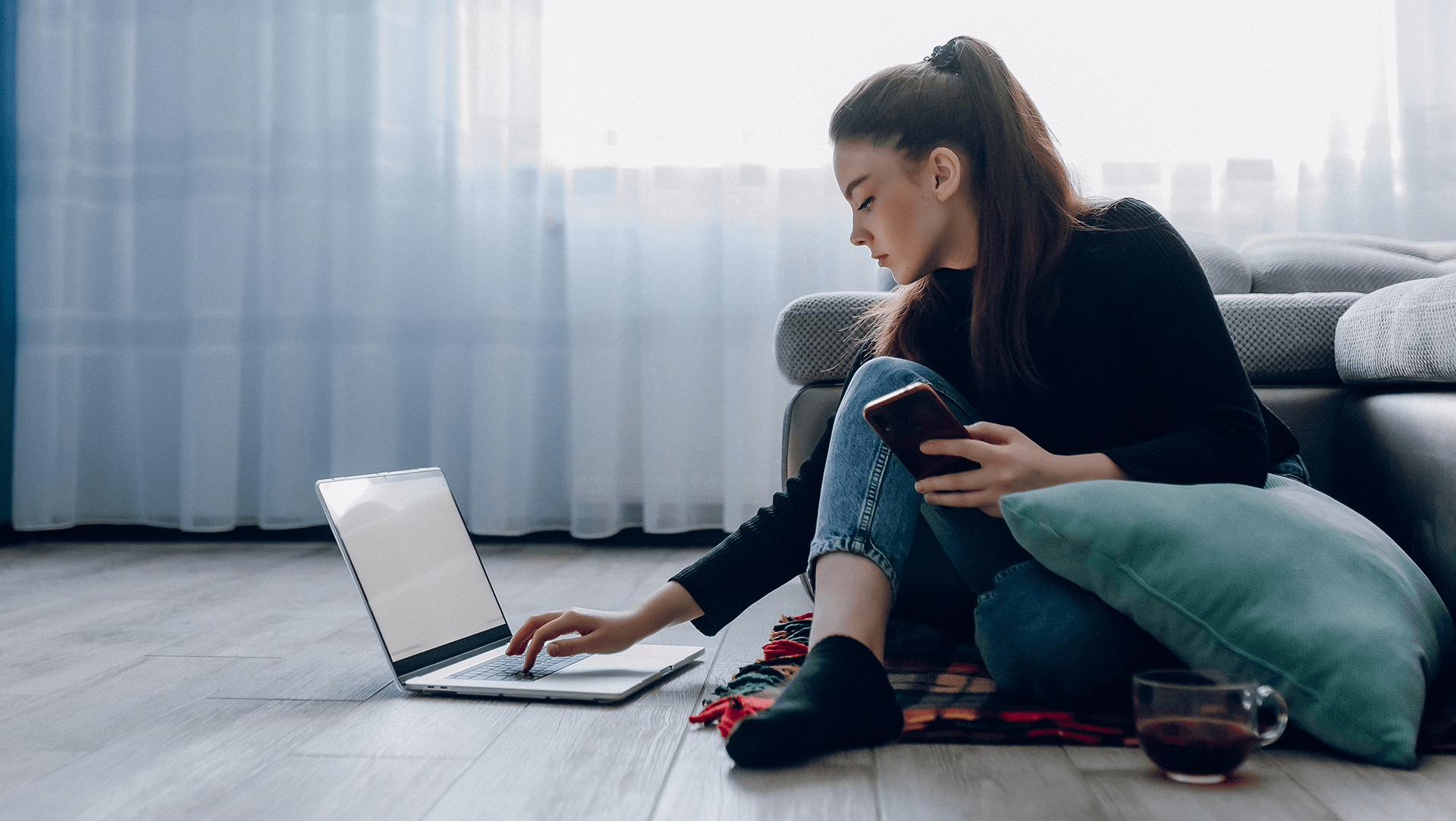 A woman sitting on the floor leaning against a couch, working on a laptop with a smartphone in one hand and a glass of wine nearby.