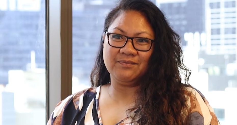 A smiling woman with long hair wearing glasses and a patterned blouse stands indoors, with a backdrop of windows revealing a high-rise urban setting.