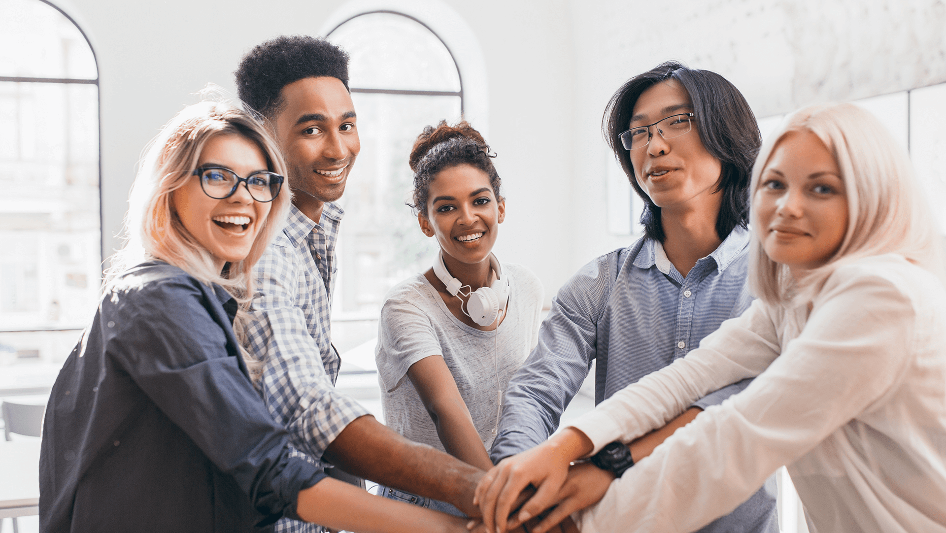 A diverse team of young professionals stacking hands in a gesture of unity and teamwork in a bright office space.