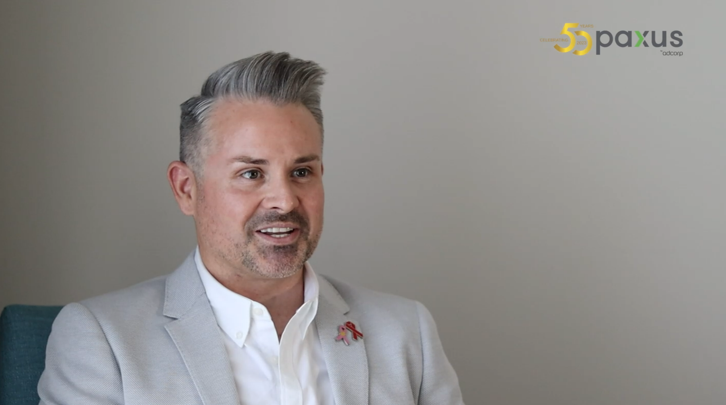 A smartly dressed person with a styled haircut, wearing a light-colored suit jacket and a red ribbon pin, engaged in a conversation or interview in a room with a neutral background.