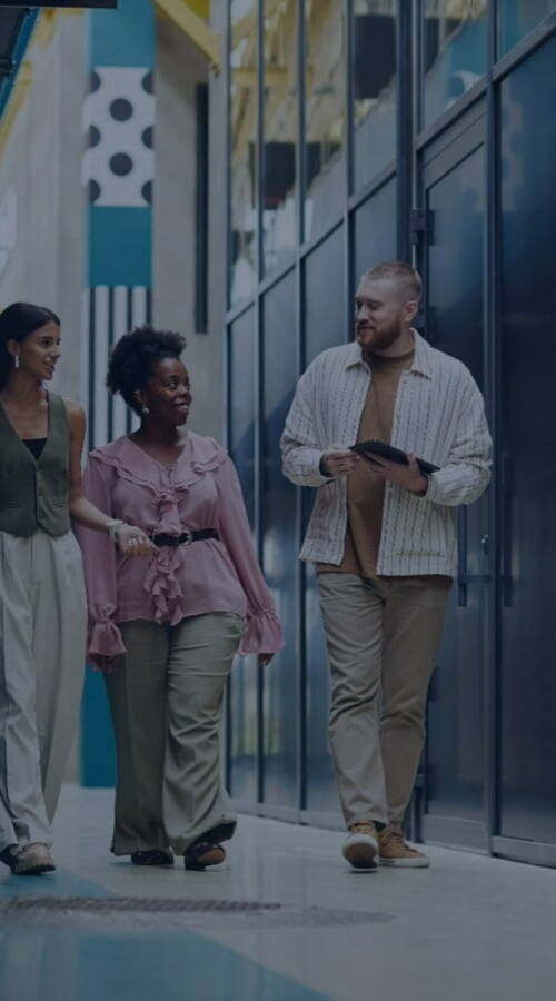 Three people walking in a modern office hallway, talking and smiling. One holds a tablet. The corridor features glass walls and a sleek design.