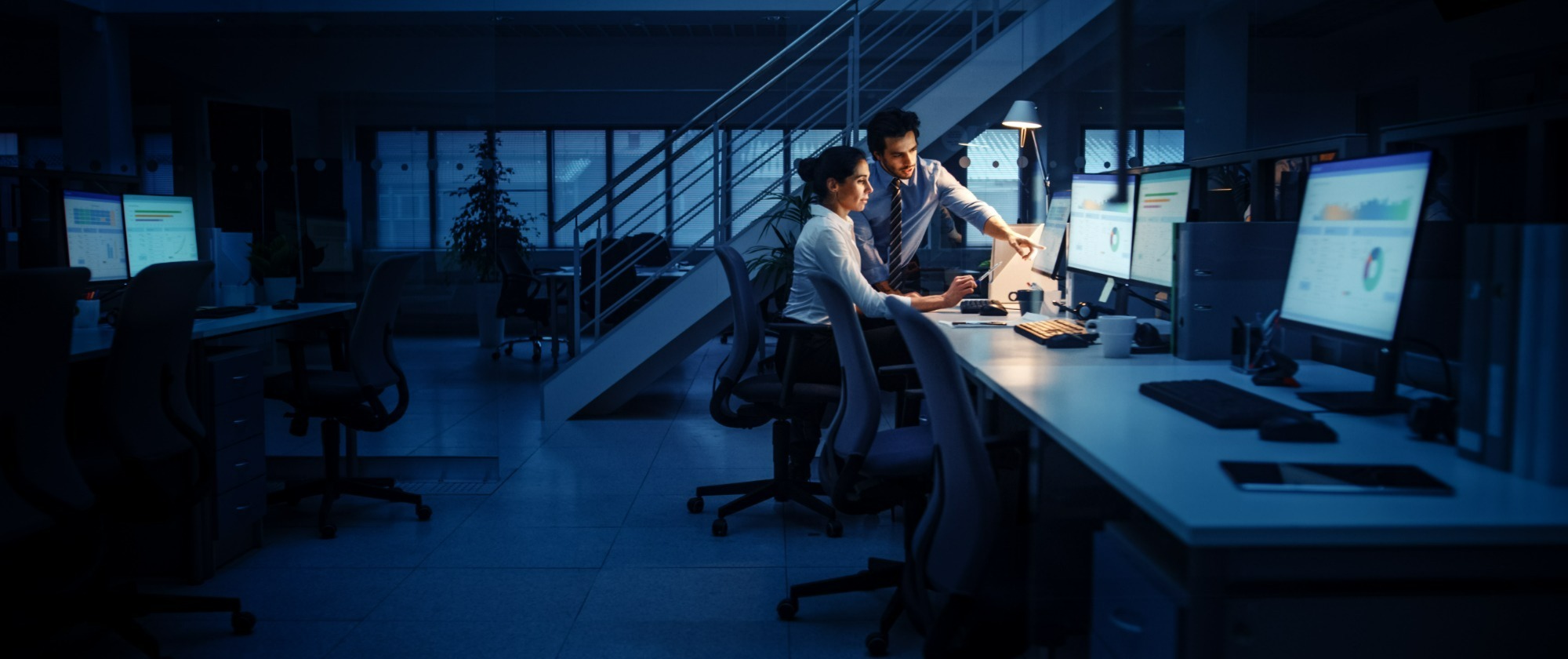 Two people working late in a dimly-lit office, seated at a desk with multiple computer screens displaying charts and graphs. One person is sitting, looking at the screen, while the other stands beside them, pointing at the monitor, discussing something.