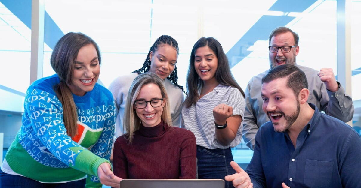 A group of cheerful colleagues celebrating a success around a laptop in a modern office setting.