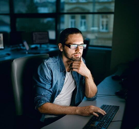 A man with a beard and glasses is seated at a desk, looking at a computer screen. He is wearing a denim shirt over a white T-shirt, working in a dimly lit room with large windows in the background.