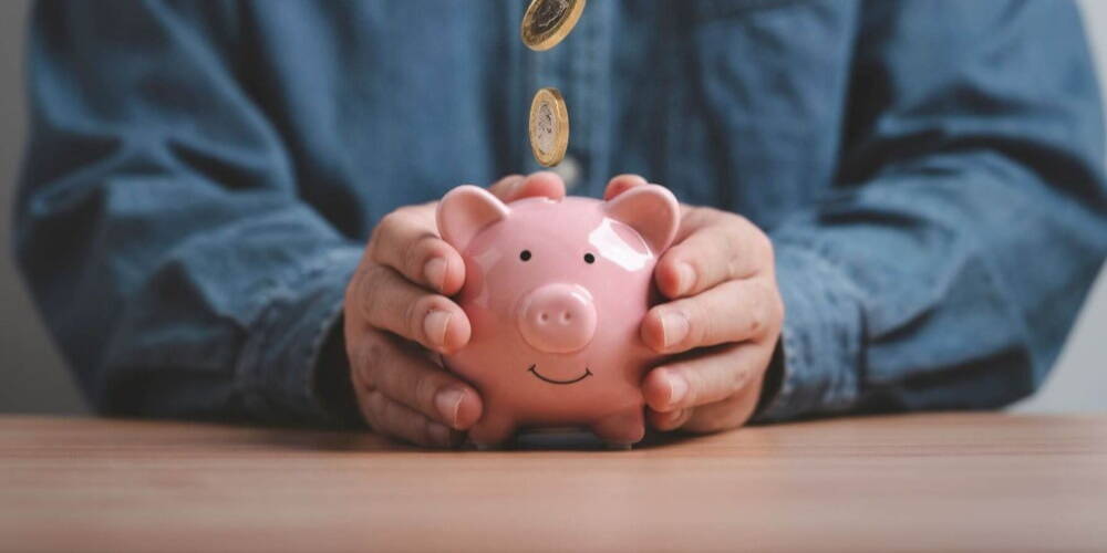 A person in a denim shirt saving money by dropping coins into a smiling piggy bank.