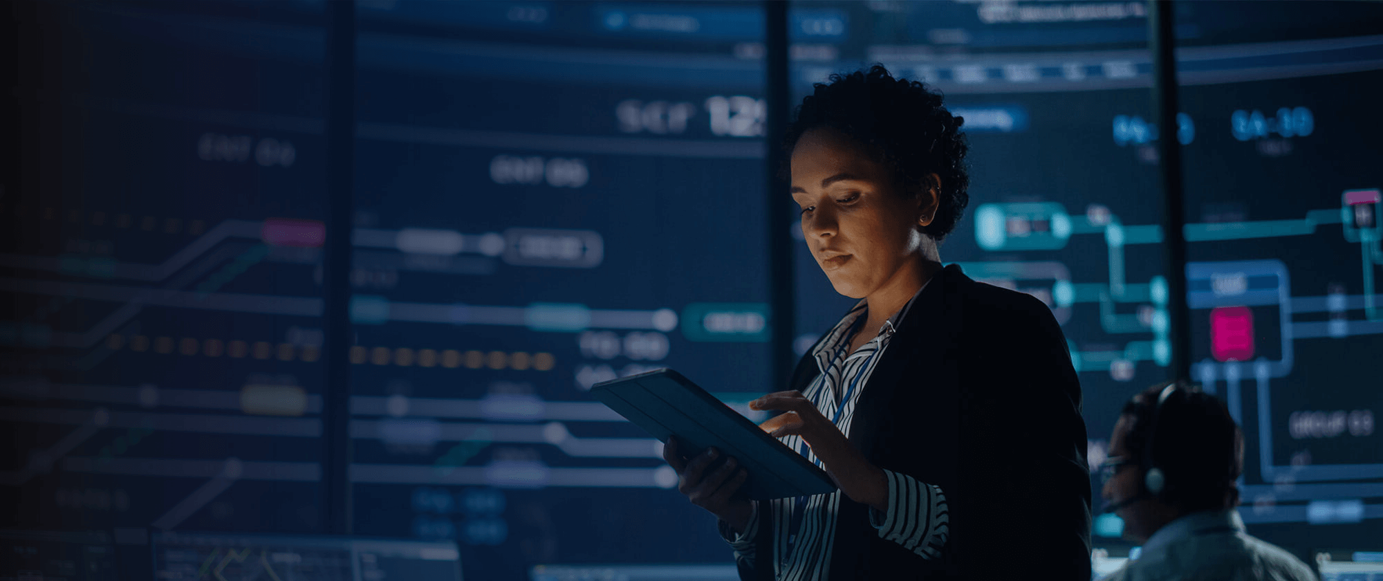 A focused professional analyzing data on a tablet with interactive analytics displayed on monitors in a high-tech control room.