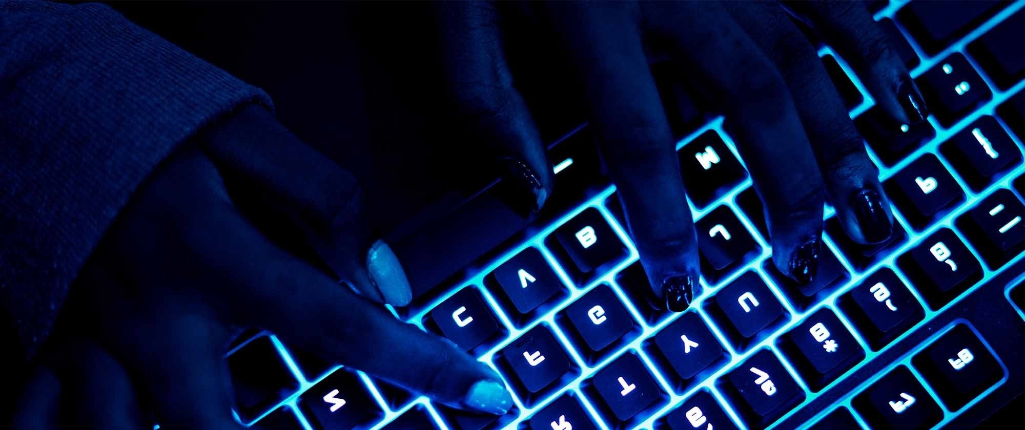 Hands typing on a backlit keyboard in a dimly lit environment, highlighting the interplay of technology and user interaction.