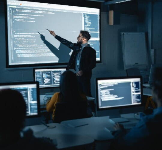 A person is presenting code on a projector screen in a dimly lit classroom. Several students are seated at desks with laptops, following along.