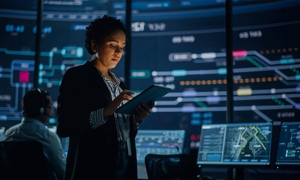 A focused professional analyzing data on a tablet with interactive analytics displayed on monitors in a high-tech control room.