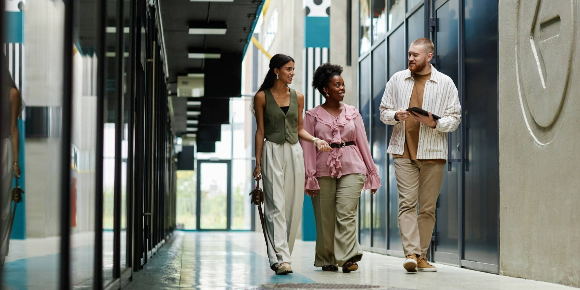 Three people walking in a modern office hallway, talking and smiling. One holds a tablet. The corridor features glass walls and a sleek design.