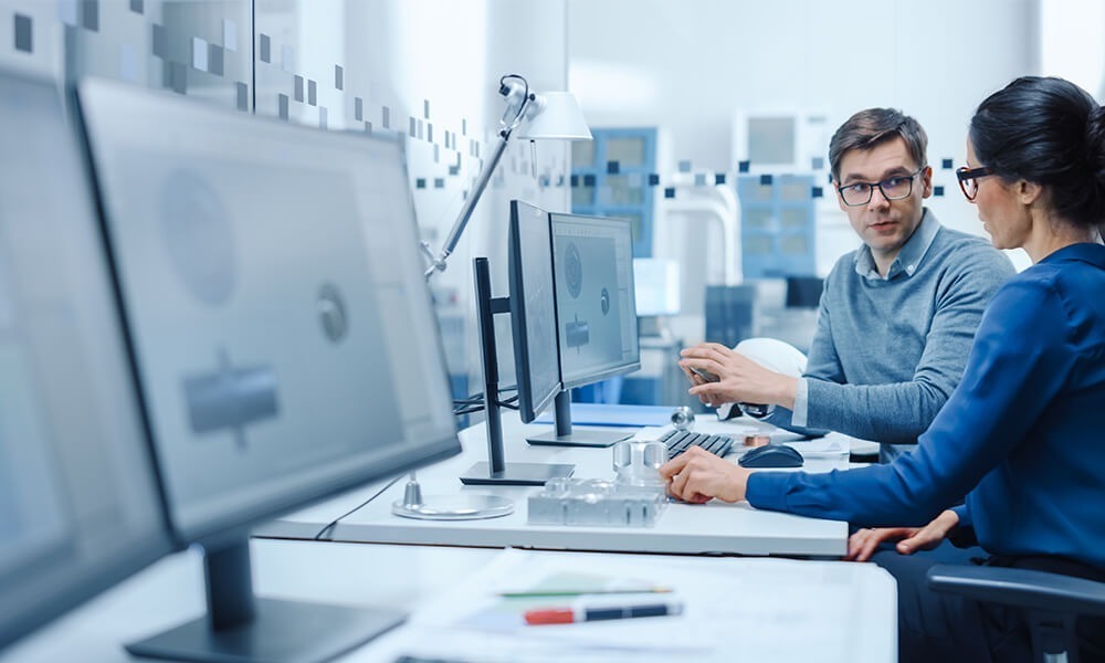 Two professionals discussing work at a computer workstation in a modern office.